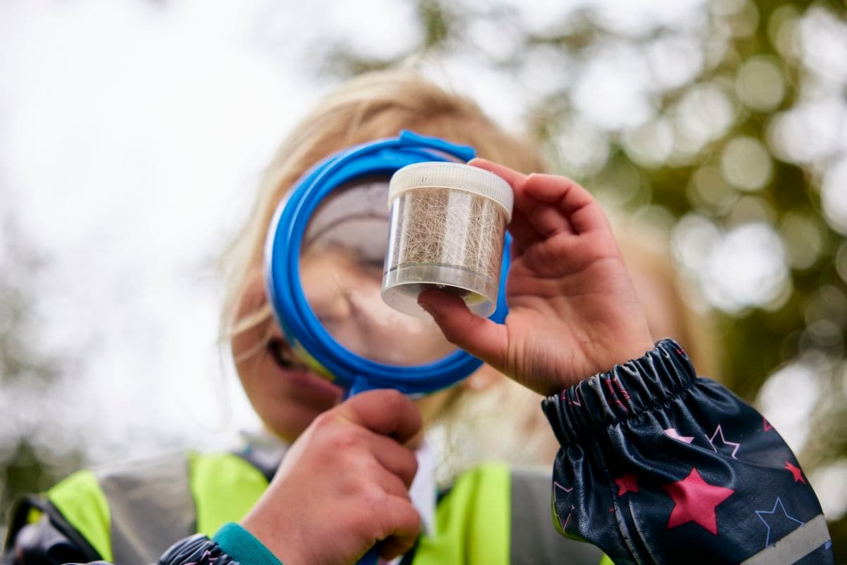 A close up of a young girl looking through a magnifying glass at a pot of dried grass