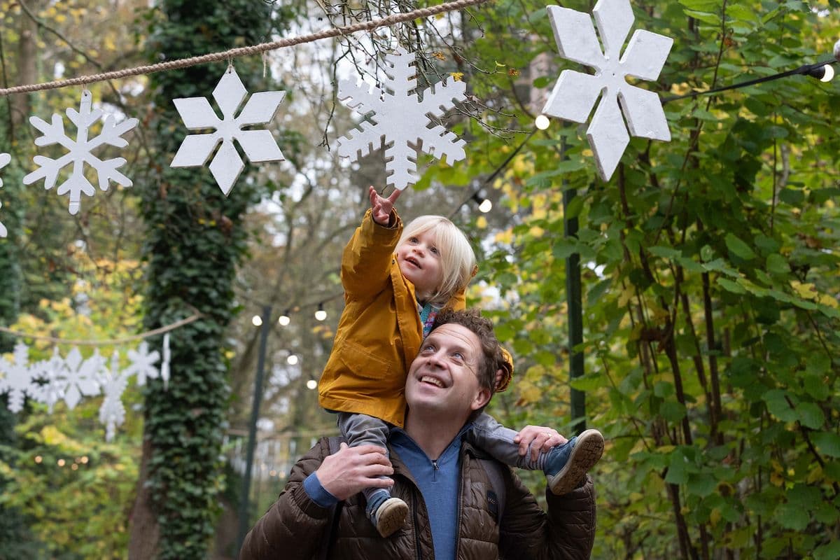 Dad with daughter on his shoulders, walking through a Festive Woodland. Daughter reaches to touch a snowflake hanging from the trees.